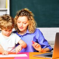 Female elementary school teacher helping little boy with writing lesson in classroom at elementary school. Back to school. Learning and education concept. Mother and little son schooling together.